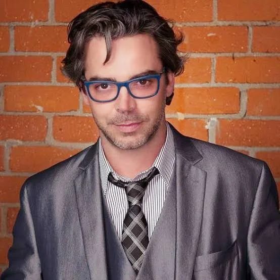 Will Keenan in a grey suit and blue glasses against a brick wall